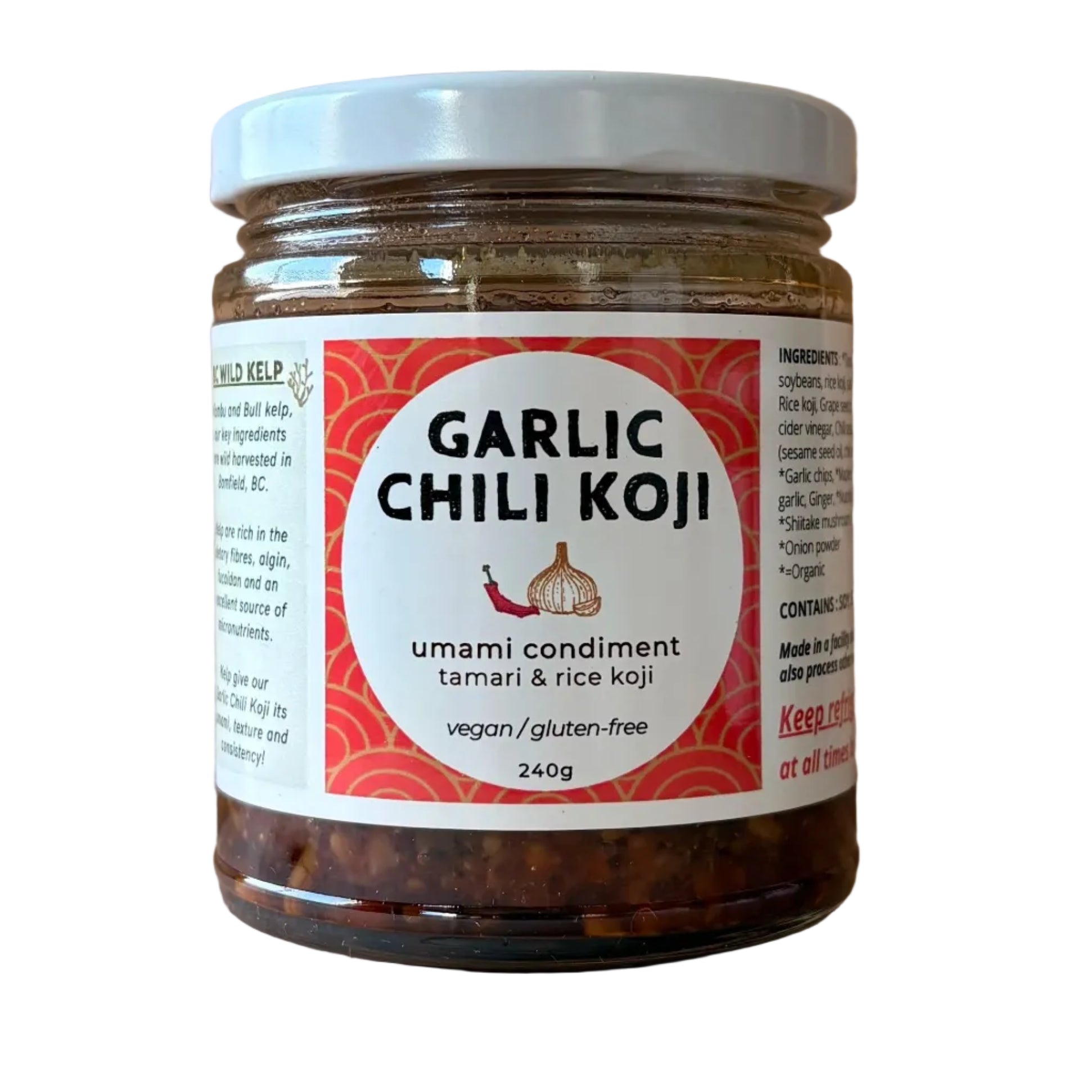 A jar of Garlic Chili Koji umami condiment on a light wood background. The clear glass jar shows the dark red chili paste inside, topped with a white lid and a white label with red and black text.