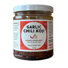 A jar of Garlic Chili Koji umami condiment on a light wood background. The clear glass jar shows the dark red chili paste inside, topped with a white lid and a white label with red and black text.