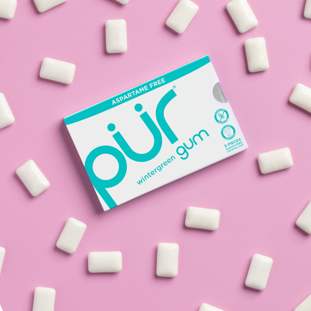 Box of PUR wintergreen gum on a pink background with gum pieces around